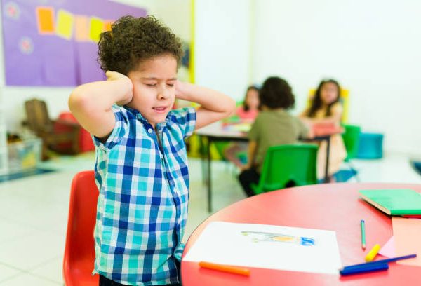 Upset autistic little boy covering his ears and feeling distressed and overwhelmed by the loud noises in preschool